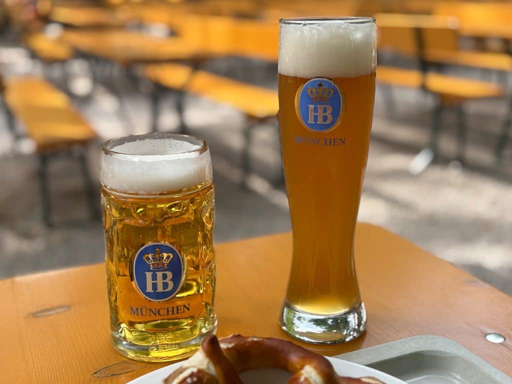 Zwei Gläser Hofbräu-Bier, ein Maßkrug und ein Weißbierglas, stehen neben einer Brezel auf einem Holztisch im Biergarten Taxisgarten in München.