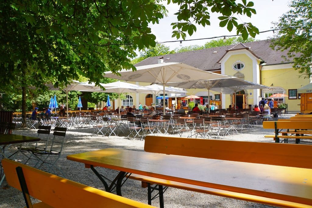 Ein sonniger Blick auf den Taxisgarten Biergarten in München mit zahlreichen Holztischen und Bänken unter großen weißen Sonnenschirmen und schattenspendenden Bäumen vor einem gelben Traditionsgebäude.