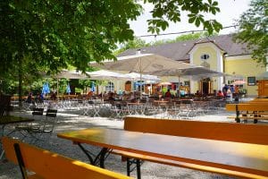 Ein sonniger Blick auf den Taxisgarten Biergarten in München mit zahlreichen Holztischen und Bänken unter großen weißen Sonnenschirmen und schattenspendenden Bäumen vor einem gelben Traditionsgebäude.