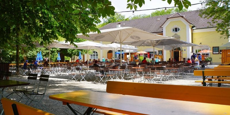 Ein sonniger Blick auf den Taxisgarten Biergarten in München mit zahlreichen Holztischen und Bänken unter großen weißen Sonnenschirmen und schattenspendenden Bäumen vor einem gelben Traditionsgebäude.