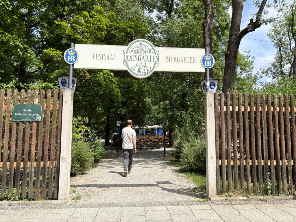 Eingang zum Biergarten Taxisgarten in München mit einem großen weißen Schild über dem Weg, flankiert von Holzzäunen und schattigen Bäumen. Ein Mann geht durch den Eingangsbereich in Richtung der im Hintergrund sichtbaren Biertische.
