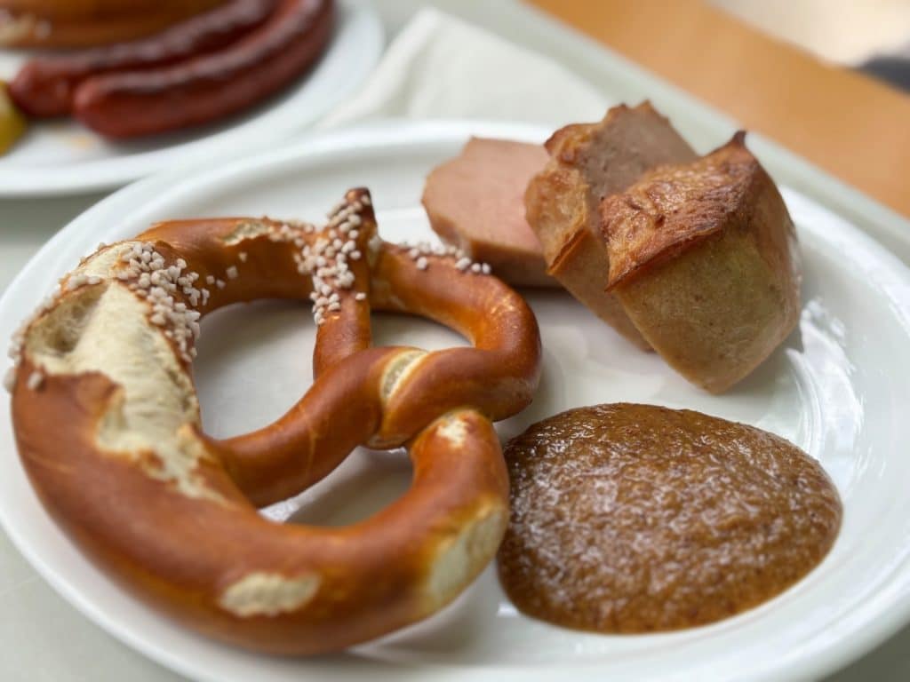 Ein weißer Teller mit einer bayerischen Brotzeit aus dem Münchner Taxisgarten, bestehend aus einer mit grobem Salz bestreuten Brezel, zwei Scheiben Leberkäse und einer Portion süßem Senf.