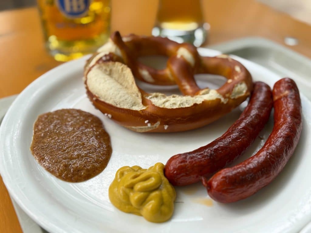 Ein weißer Teller im Taxisgarten in München, angerichtet mit zwei gebratenen Würsten, einer großen Brezel sowie Portionen von süßem und mittelscharfem Senf vor einem Glas Bier.