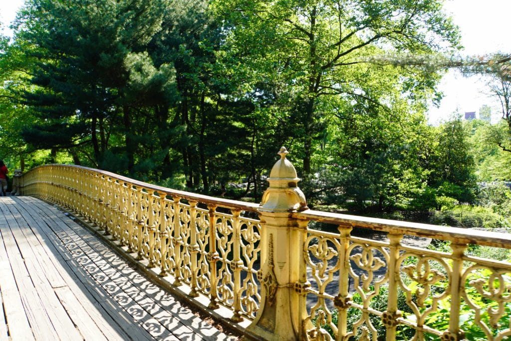 Eine geschwungene Brücke mit einem verzierten, hellgelben gusseisernen Geländer und Holzplanken führt im New Yorker Central Park durch eine dichte, grüne Parklandschaft.