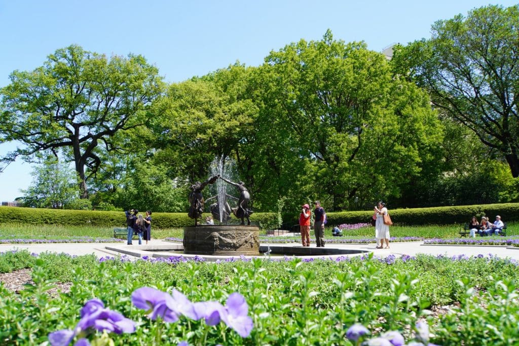 Der Untermyer-Brunnen mit seinen tanzenden Bronzefiguren steht im Zentrum des Conservatory Garden im Central Park von New York, umgeben von blühenden lila Blumenbeeten, grünen Hecken und hohen Bäumen unter blauem Himmel.