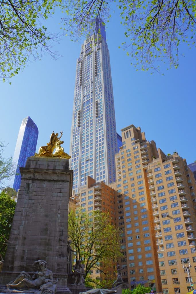 Blick vom Central Park in New York auf das vergoldete Maine-Monument im Vordergrund, hinter dem sich die moderne Skyline mit dem markanten Wolkenkratzer 220 Central Park South unter blauem Himmel erhebt.