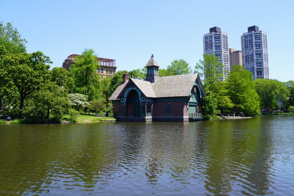 Ein historisches Bootshaus mit Backsteinfassade und spitzem Turm steht am Ufer eines Sees im Central Park in New York, umrahmt von grünen Bäumen und modernen Hochhäusern im Hintergrund.