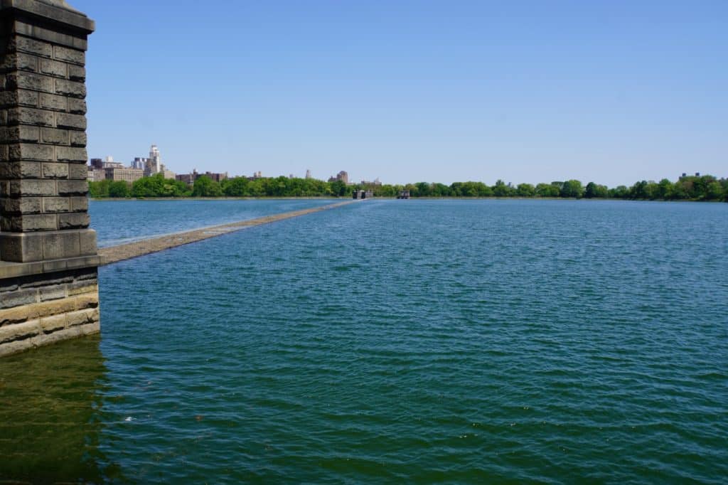 Blick über das weite, blaue Wasser des Jacqueline Kennedy Onassis Reservoirs im Central Park in New York, mit einer steinernen Ufermauer im Vordergrund und der fernen Skyline hinter einem dichten Grüngürtel.