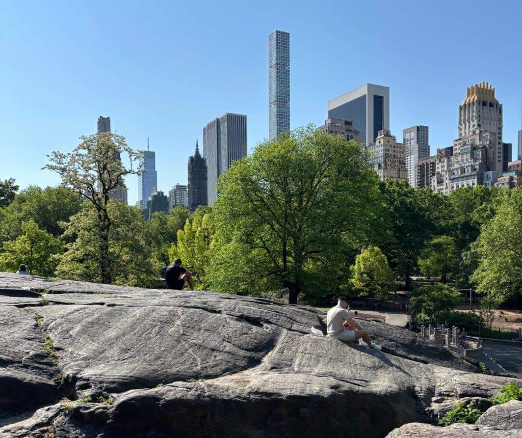 Blick vom Umpire Rock im New York Central Park auf Menschen, die auf den Felsen entspannen, mit der markanten Skyline von Manhattan und dem Central Park Tower im Hintergrund.
