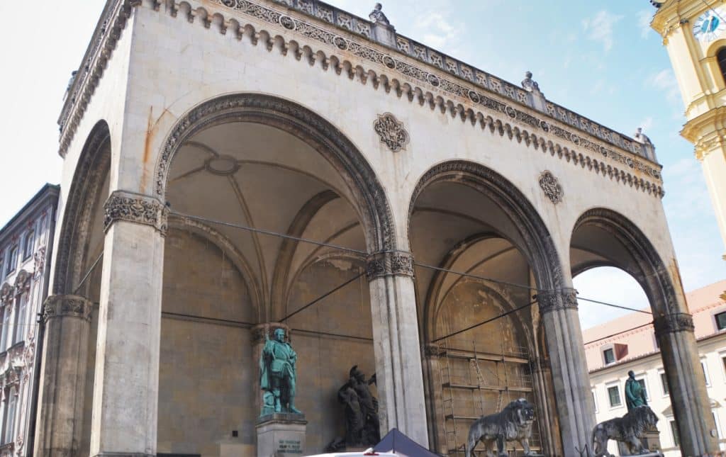 Die Feldherrnhalle in München präsentiert sich als monumentale, offene Loggia mit drei hohen Rundbögen, unter denen Bronzestatuen und steinerne Löwen vor einer hellen Steinfassade thronen.
