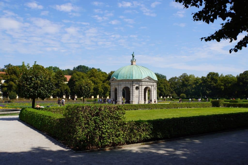 Der Dianatempel im Münchner Hofgarten steht zentral in einer gepflegten Parkanlage mit grünen Hecken und Rasenflächen unter einem leicht bewölkten blauen Himmel.