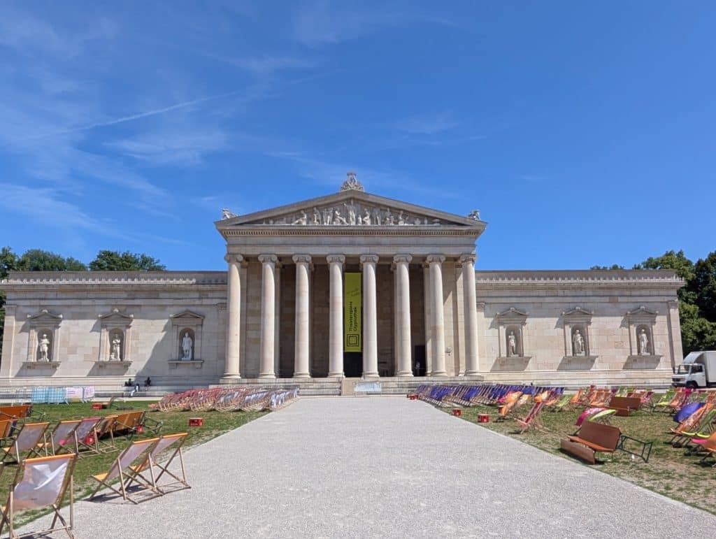 Die Glyptothek am Königsplatz in München zeigt sich als klassizistisches Gebäude mit imposanter Säulenfront und Skulpturen in Nischen, davor erstreckt sich ein weiter Platz mit zahlreichen bunten Liegestühlen unter blauem Himmel.