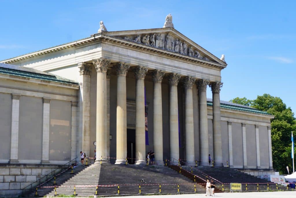 Die Glyptothek am Königsplatz in München zeigt sich als klassizistisches Gebäude mit einer monumentalen Säulenfront, einem figurengeschmückten Giebel und einer breiten Freitreppe unter blauem Himmel.