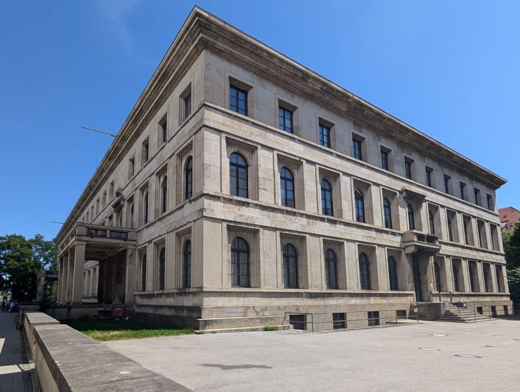 Ein monumentaler, dreistöckiger Steinbau aus der Zeit des Nationalsozialismus in München mit einer strengen Fassade, hohen Rundbogenfenstern und einem massiven Säulenportal unter strahlend blauem Himmel.