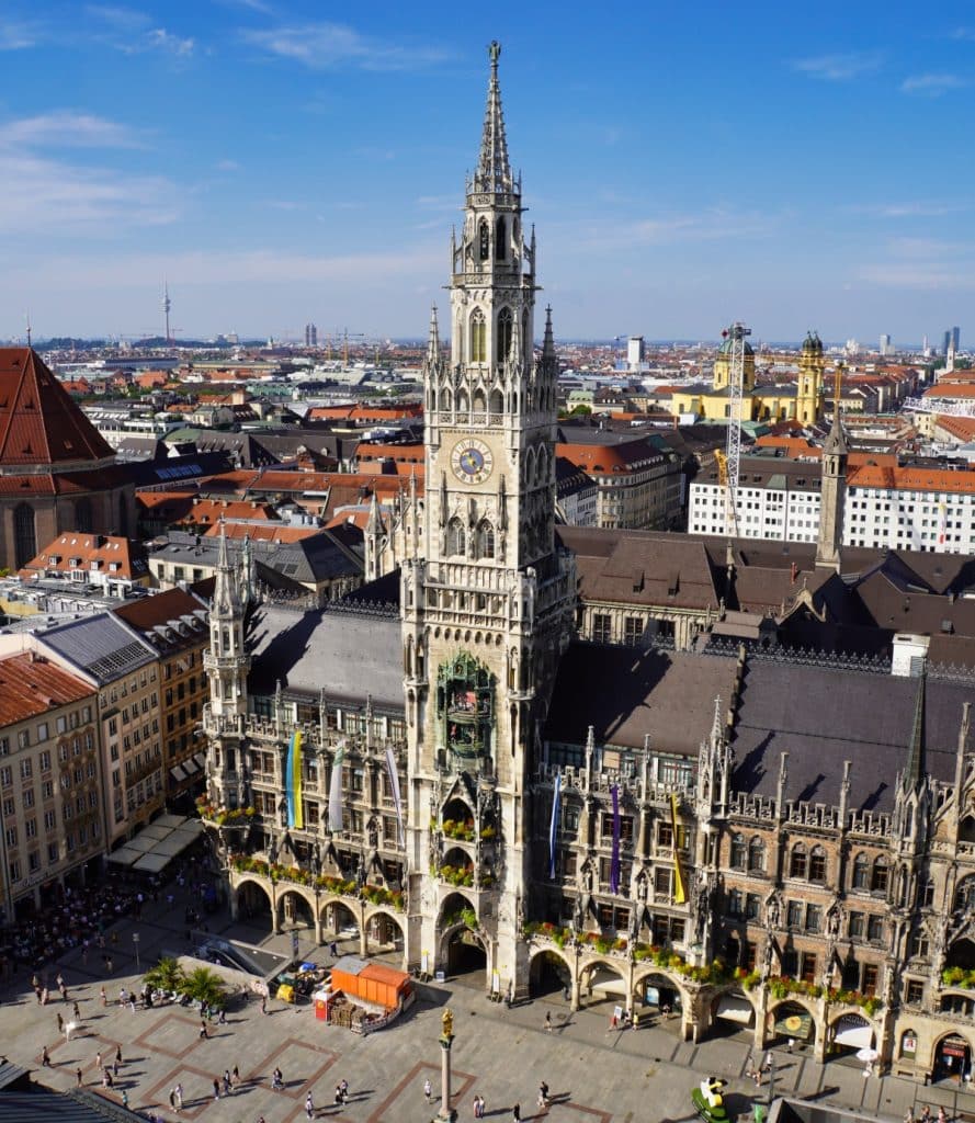 Luftaufnahme des Neuen Rathauses in München am Marienplatz mit seiner neugotischen Fassade, dem markanten Glockenspielturm und dem belebten Platz davor unter blauem Himmel.