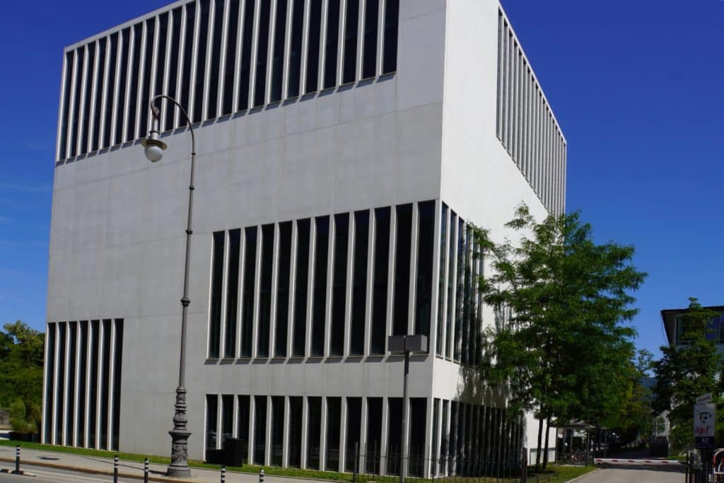 Das NS-Dokumentationszentrum in München präsentiert sich als moderner, würfelförmiger Bau aus weißem Sichtbeton mit markanten, vertikalen Fensterbändern vor einem strahlend blauen Himmel.
