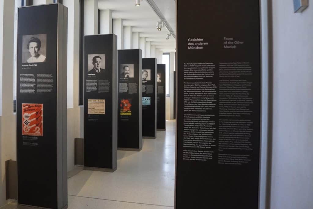 Eine Reihe von hohen, schwarzen Informationstafeln in der Ausstellung des NS-Dokumentationszentrums München zeigt Porträts, Texte und historische Dokumente unter dem Titel „Gesichter des anderen München“. Die Stelen stehen in einem hellen, modern gestalteten Raum mit großen Fenstern und beleuchten den Widerstand gegen den Nationalsozialismus.