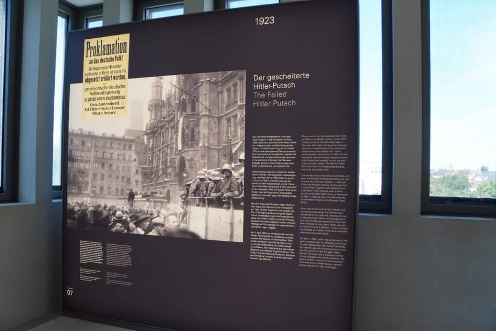 Eine Informationstafel im NS-Dokumentationszentrum München zeigt unter der Überschrift „1923 Der gescheiterte Hitler-Putsch“ historische Fotografien und Begleittexte zum versuchten Staatsstreich. Das zentrale Bild zeigt bewaffnete Putschisten auf einem Lastwagen vor dem Münchner Rathaus, ergänzt durch das Faksimile einer Proklamation der „provisorischen deutschen Nationalregierung“.