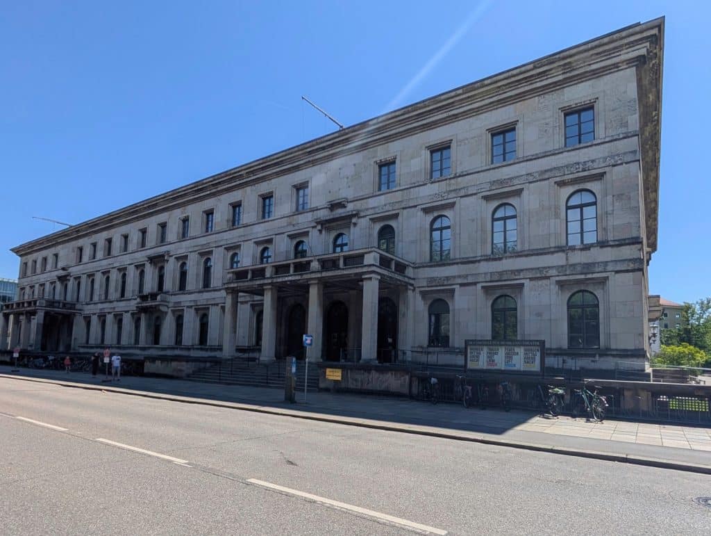Das Bild zeigt das monumentale, neoklassizistische Gebäude der ehemaligen NSDAP-Parteizentrale in München an einem sonnigen Tag unter blauem Himmel. Die langgestreckte Steinfassade mit ihren gleichmäßigen Fensterreihen und dem markanten Säulenportal erstreckt sich entlang einer breiten Straße.