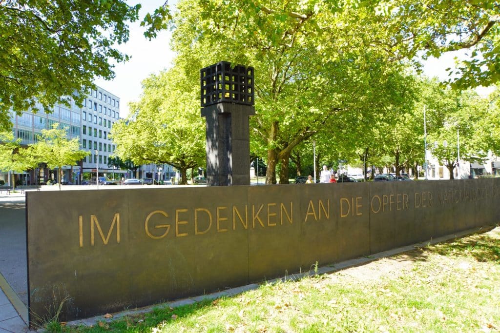 Das Mahnmal für die Opfer der Nationalsozialisten am Platz der Opfer des Nationalsozialismus in München zeigt eine dunkle Steinmauer mit der goldenen Inschrift „IM GEDENKEN AN DIE OPFER DER NATIONALSOZIALISTISCHEN GEWALTHERRSCHAFT“ vor einer ewigen Flamme in einem Metallkäfig. Im Hintergrund sind grüne Bäume und städtische Gebäude unter einem hellen Himmel zu sehen.