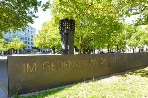 Das Mahnmal für die Opfer der Nationalsozialisten am Platz der Opfer des Nationalsozialismus in München zeigt eine dunkle Steinmauer mit der goldenen Inschrift „IM GEDENKEN AN DIE OPFER DER NATIONALSOZIALISTISCHEN GEWALTHERRSCHAFT“ vor einer ewigen Flamme in einem Metallkäfig. Im Hintergrund sind grüne Bäume und städtische Gebäude unter einem hellen Himmel zu sehen.