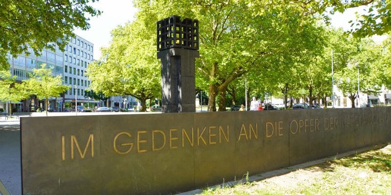 Das Mahnmal für die Opfer der Nationalsozialisten am Platz der Opfer des Nationalsozialismus in München zeigt eine dunkle Steinmauer mit der goldenen Inschrift „IM GEDENKEN AN DIE OPFER DER NATIONALSOZIALISTISCHEN GEWALTHERRSCHAFT“ vor einer ewigen Flamme in einem Metallkäfig. Im Hintergrund sind grüne Bäume und städtische Gebäude unter einem hellen Himmel zu sehen.