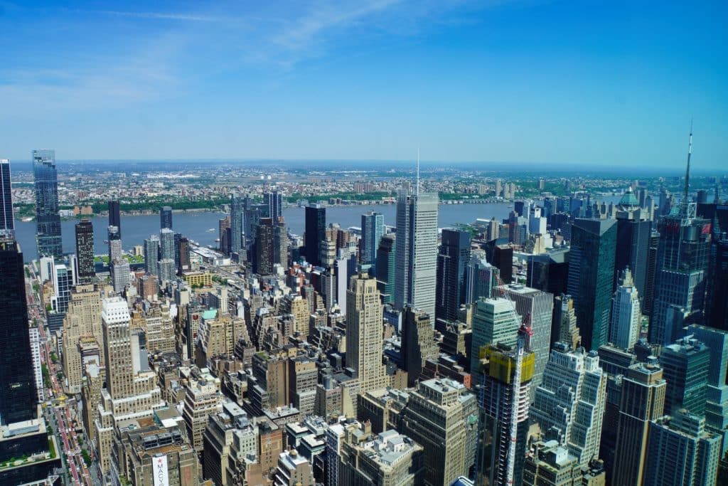 Blick vom Empire State Building über die dicht bebaute Skyline von Manhattan mit zahlreichen Wolkenkratzern, dem Hudson River und New Jersey im Hintergrund unter einem klaren blauen Himmel.
