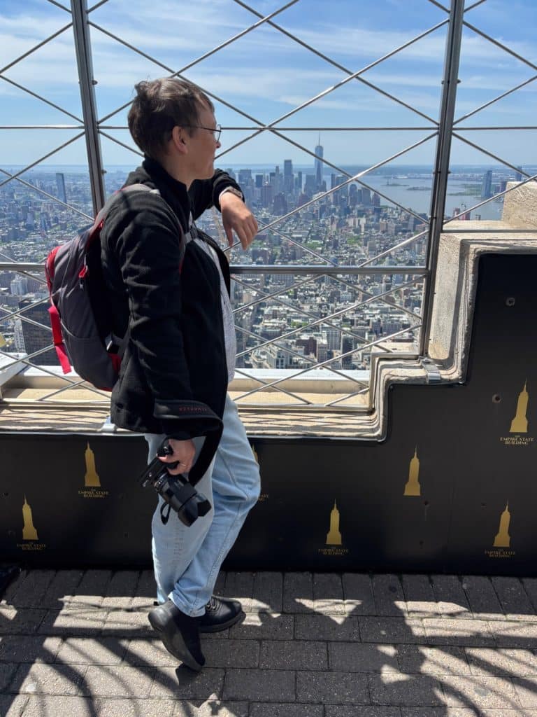Eine Person mit Brille, Rucksack und Kamera in der Hand blickt von der Aussichtsplattform des Empire State Buildings über die Skyline von New York City. Im Hintergrund erstreckt sich das Stadtpanorama mit Wolkenkratzern und dem Hudson River unter einem klaren blauen Himmel.