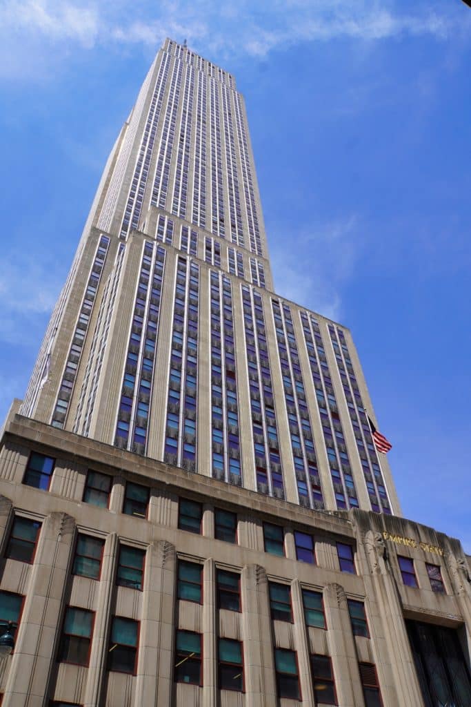 Eine Untersicht des Empire State Building in New York, dessen Art-déco-Fassade aus Kalkstein und zahlreichen Fensterreihen sich vor einem strahlend blauen Himmel in die Höhe streckt. Über dem Eingangsbereich weht eine amerikanische Flagge an der Seite des monumentalen Wolkenkratzers.