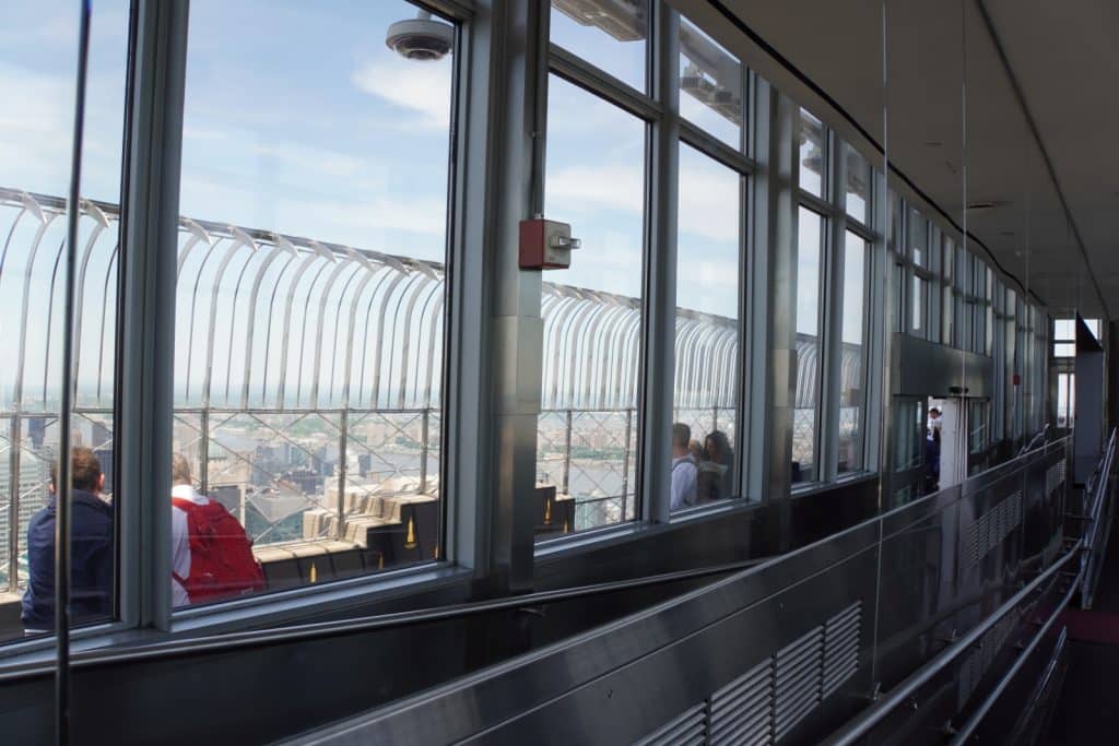 Blick von der verglasten Aussichtsplattform des Empire State Building in New York auf die Stadt, mit Besuchern im Vordergrund und dem charakteristischen Metallzaun vor den Fenstern.