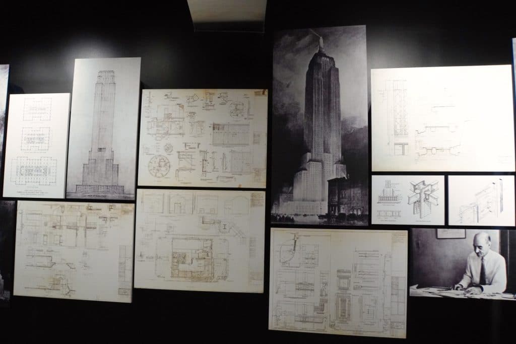 Eine Wanderausstellung zeigt historische Baupläne, technische Zeichnungen und Skizzen des Empire State Building in New York sowie ein Schwarz-Weiß-Foto eines Architekten bei der Arbeit.