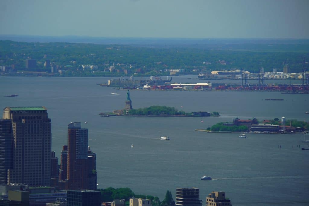 Weitwinkel-Blick vom Empire State Building auf den New Yorker Hafen mit der Freiheitsstatue auf Liberty Island im Zentrum, umgeben von Wasserstraßen und der fernen Küstenlinie. Im Vordergrund sind Teile der Skyline von Lower Manhattan und vorbeifahrende Schiffe auf dem Hudson River zu sehen.