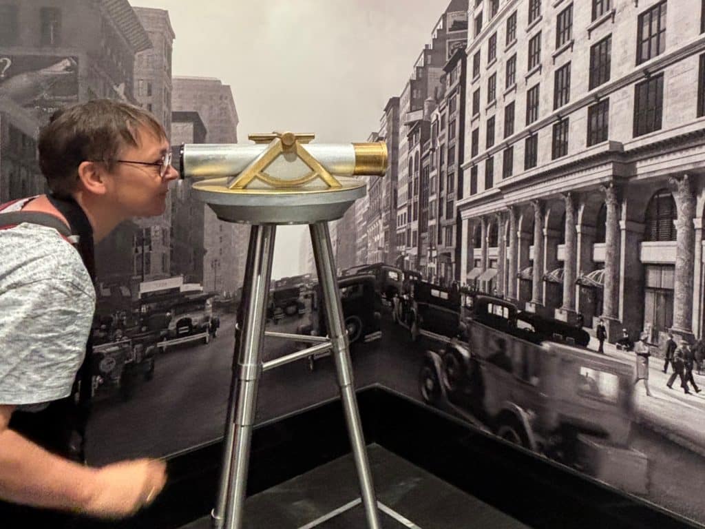 Eine Person blickt durch ein historisches Vermessungsgerät auf eine großformatige Schwarz-Weiß-Fotografie im Empire State Building, die eine belebte Straßenszene im historischen New York zeigt.