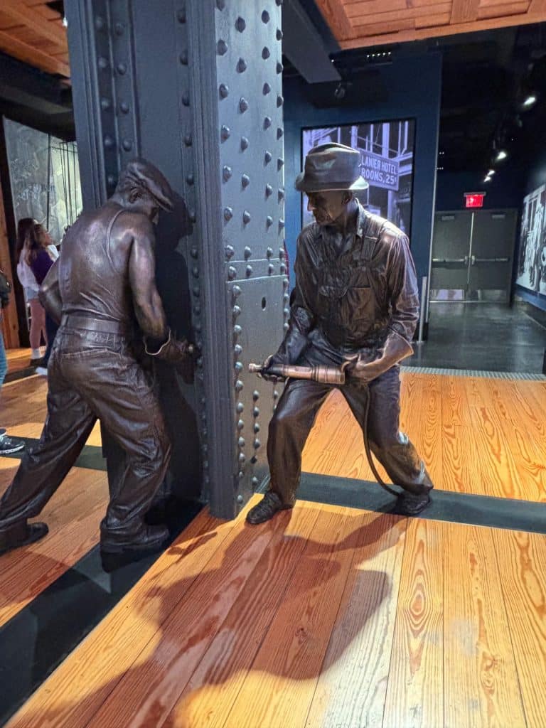 Zwei lebensgroße Bronzestatuen von Arbeitern am Empire State Building stellen den Bauprozess nach, wobei einer eine schwere Stahlsäule stützt und der andere ein Nietwerkzeug bedient.