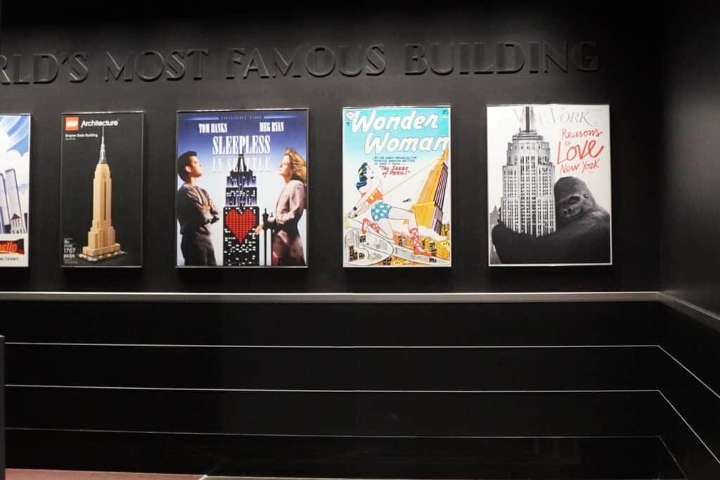 Eine schwarze Wand mit der Aufschrift „WORLD'S MOST FAMOUS BUILDING“ zeigt verschiedene Plakate, die das Empire State Building in New York in der Popkultur darstellen, darunter Motive von LEGO, „Sleepless in Seattle“, Wonder Woman und King Kong.