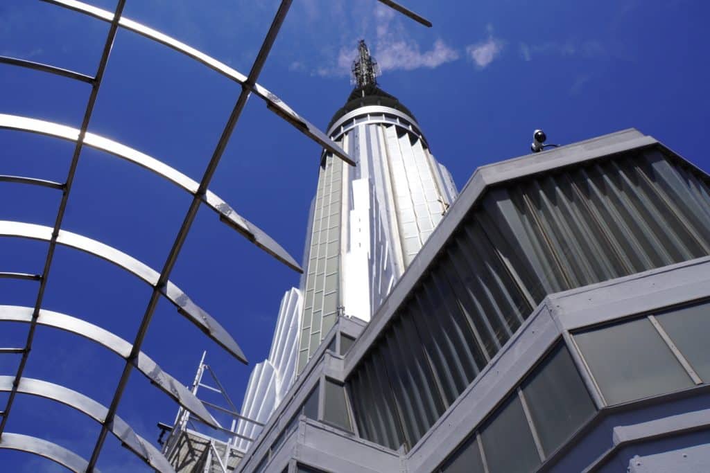 Blick von unten auf die glänzende Spitze des Empire State Building in New York gegen einen strahlend blauen Himmel, eingerahmt von einer metallischen Gitterstruktur im Vordergrund.