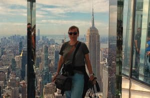 Eine Frau steht lächelnd vor einer Glaswand auf einer Aussichtsplattform in New York, während im Hintergrund das Empire State Building und die Skyline der Stadt unter einem leicht bewölkten Himmel zu sehen sind. Sie trägt eine Sonnenbrille, ein graues T-Shirt, helle Hosen und hat eine Kameratasche umgehängt.