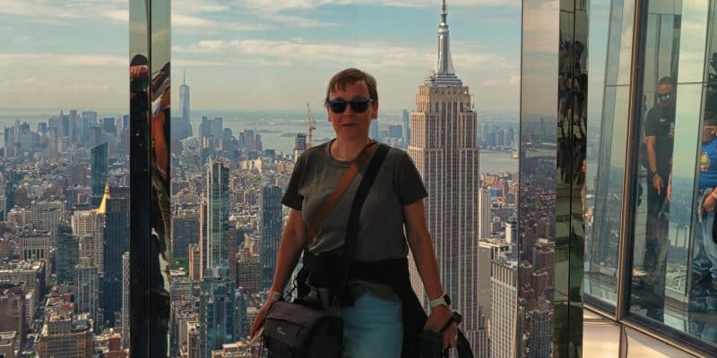Eine Frau steht lächelnd vor einer Glaswand auf einer Aussichtsplattform in New York, während im Hintergrund das Empire State Building und die Skyline der Stadt unter einem leicht bewölkten Himmel zu sehen sind. Sie trägt eine Sonnenbrille, ein graues T-Shirt, helle Hosen und hat eine Kameratasche umgehängt.