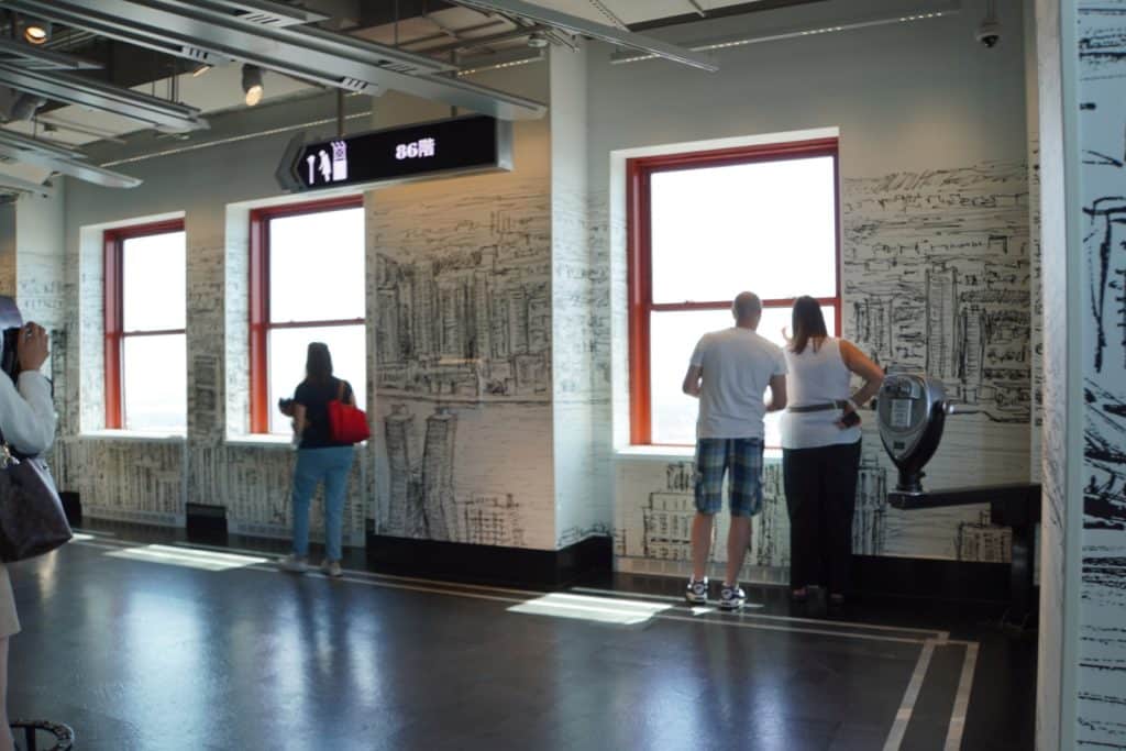 Besucher stehen auf der geschlossenen Aussichtsplattform des Empire State Building in New York und blicken durch große Fenster auf die Stadt, während die Wände mit großflächigen Stadtzeichnungen gestaltet sind. Ein Schild weist den Weg zur 86. Etage.