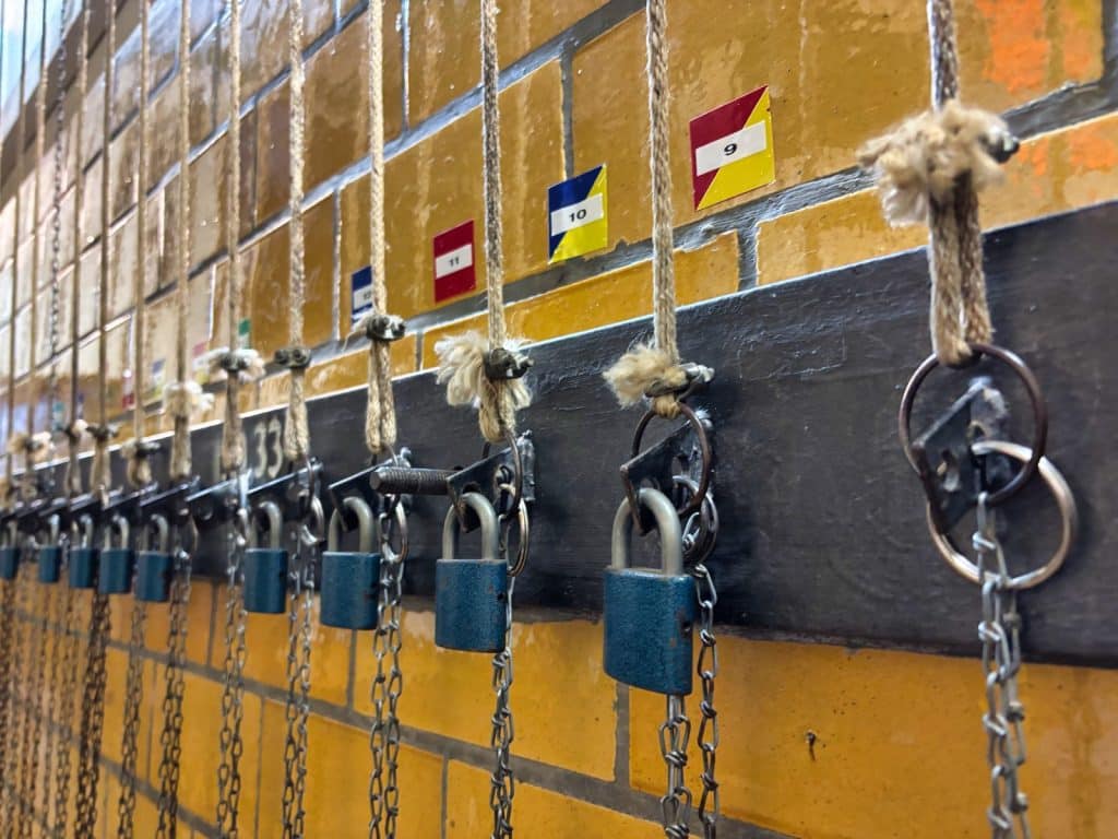 Eine Reihe von blauen Vorhängeschlössern an Ketten und Seilen sichert die Kleideraufzüge in der Kaue des Besucherbergwerks in Ehrenfriedersdorf, Sachsen. Über den Schlössern hängen nummerierte Schilder an einer gelben Ziegelwand.