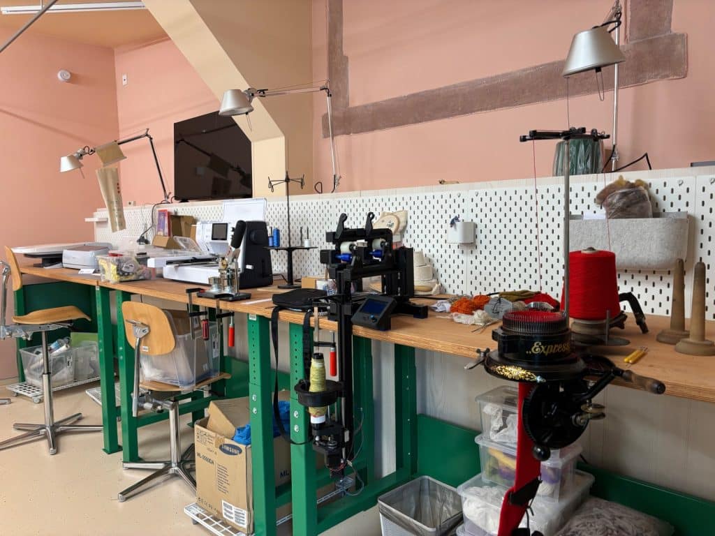 Blick in das Esche-Museum in Sachsen auf einen hellen Werkstattraum mit langen Holztischen, an denen verschiedene Näh- und Strickmaschinen sowie Garnrollen für textile Arbeiten bereitstehen.