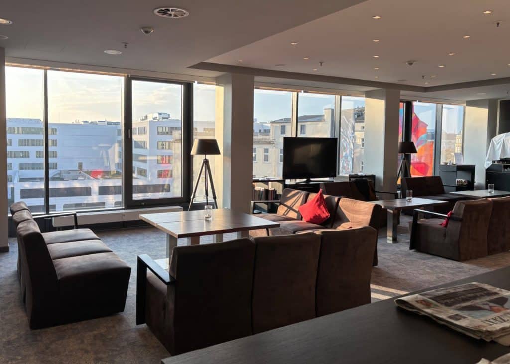Ein moderner Aufenthaltsraum im Chemnitz Hotel mit dunklen Polstersitzgruppen, Holztischen und einer Fensterfront, die den Blick auf die umliegende Stadt freigibt. Im Hintergrund befinden sich ein Fernseher und Stehlampen, während das warme Sonnenlicht den Raum flutet.