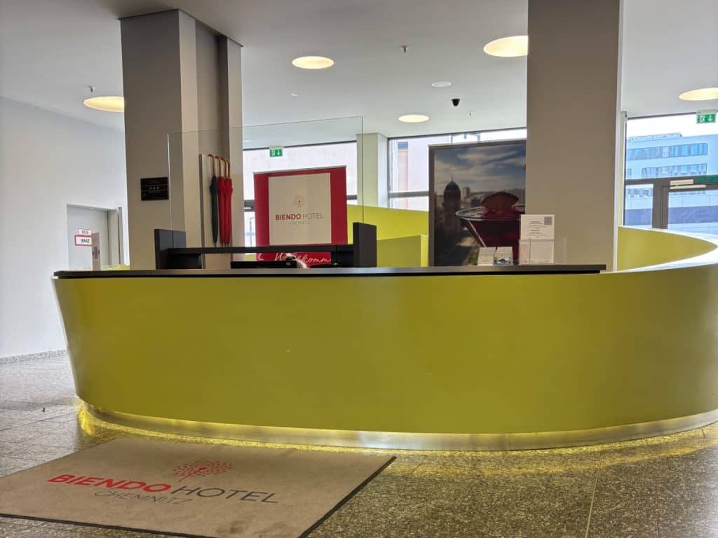 Die moderne Lobby des Biendo Hotels in Chemnitz mit einem geschwungenen, gelben Empfangstresen, einem Logo-Teppich im Vordergrund und hellen Fenstern im Hintergrund.