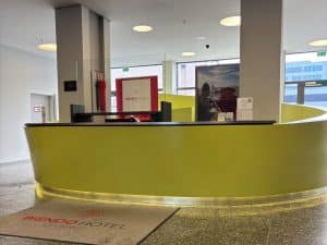 Die moderne Lobby des Biendo Hotels in Chemnitz mit einem geschwungenen, gelben Empfangstresen, einem Logo-Teppich im Vordergrund und hellen Fenstern im Hintergrund.
