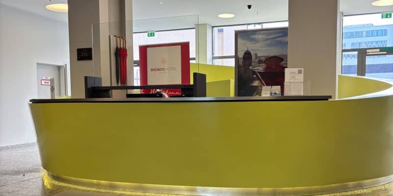 Die moderne Lobby des Biendo Hotels in Chemnitz mit einem geschwungenen, gelben Empfangstresen, einem Logo-Teppich im Vordergrund und hellen Fenstern im Hintergrund.