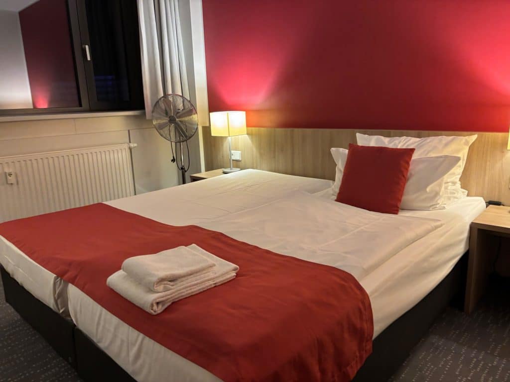 Ein modern eingerichtetes Hotelzimmer in Chemnitz mit einem Doppelbett, das mit weißer Bettwäsche, einem roten Läufer und passenden Zierkissen vor einer roten Akzentwand ausgestattet ist. Neben dem Bett befinden sich ein Nachttisch mit Lampe, ein Standventilator und ein Fenster mit hellen Vorhängen.