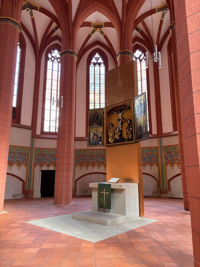 Innenansicht der Schloßkirche in Chemnitz mit Blick auf den modernen Altar und ein historisches, geöffnetes Flügelretabel vor den hohen gotischen Fenstern und roten Säulen des Chors.