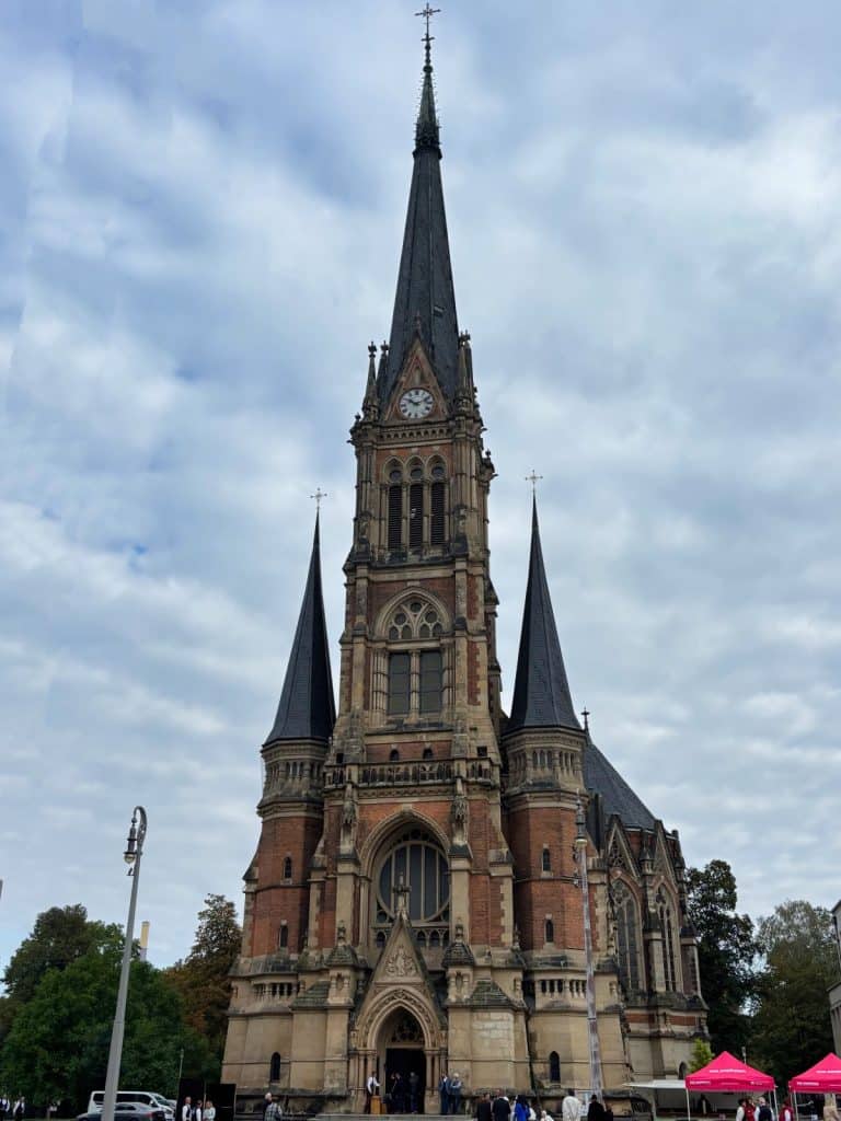 Die neugotische St.-Petri-Kirche in Chemnitz ragt mit ihrem hohen, spitzen Glockenturm und zwei kleineren Seitentürmen aus Backstein vor einem bewölkten Himmel empor. Am Fuße der Kirche auf dem Theaterplatz sind Passanten und rote Marktstände zu sehen.