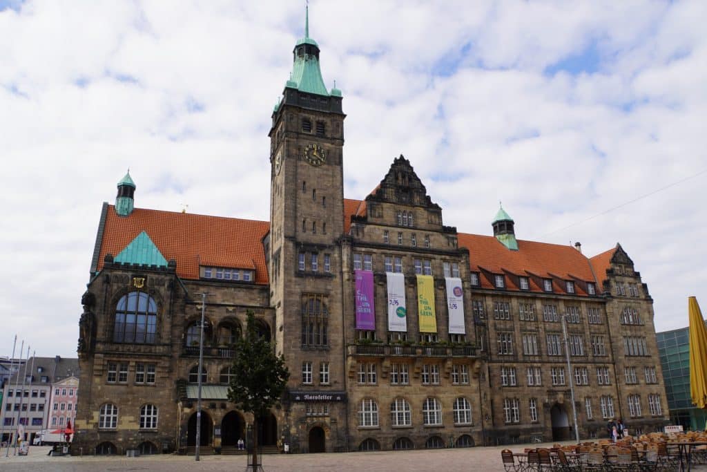 Das Neue Rathaus am Marktplatz in Chemnitz, ein monumentales Sandsteingebäude mit einem markanten Uhrturm, grünen Turmhauben und einer Fassade mit bunten Bannern. Vor dem Gebäude erstreckt sich ein gepflasterter Platz mit Außenbestuhlung eines Cafés unter einem bewölkten Himmel.