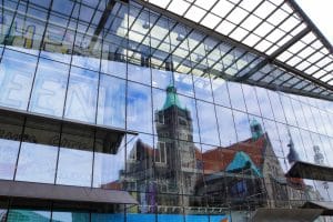In der modernen Glasfassade eines Gebäudes in Chemnitz spiegelt sich das historische Rathaus mit seinem markanten grünen Turm und den roten Ziegeldächern unter einem leicht bewölkten Himmel.